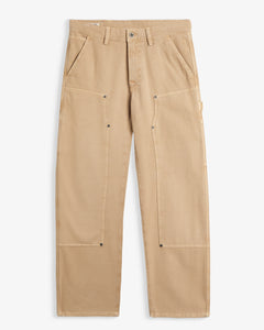 A front view of The Carpenter Pant in beige, laid flat on a white background. The image highlights the double knee panels with copper-toned rivets, slash pockets, and the embroidered c'est normal logo.