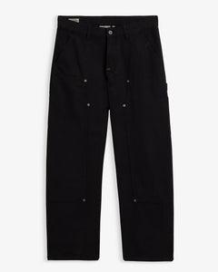 A front view of The Carpenter Pant in black, laid flat on a white background. The image highlights the double-knee panels, copper-toned rivets, slash pockets, and the embroidered c'est normal logo on the side seam.