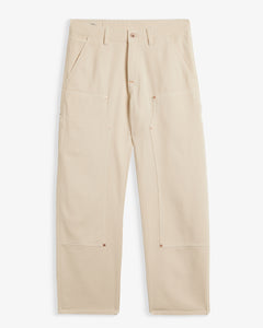A front view of The Carpenter Pant in sand, laid flat on a white background. The image highlights the double-knee panels, copper-toned rivets, and the embroidered c'est normal logo by the left pocket.