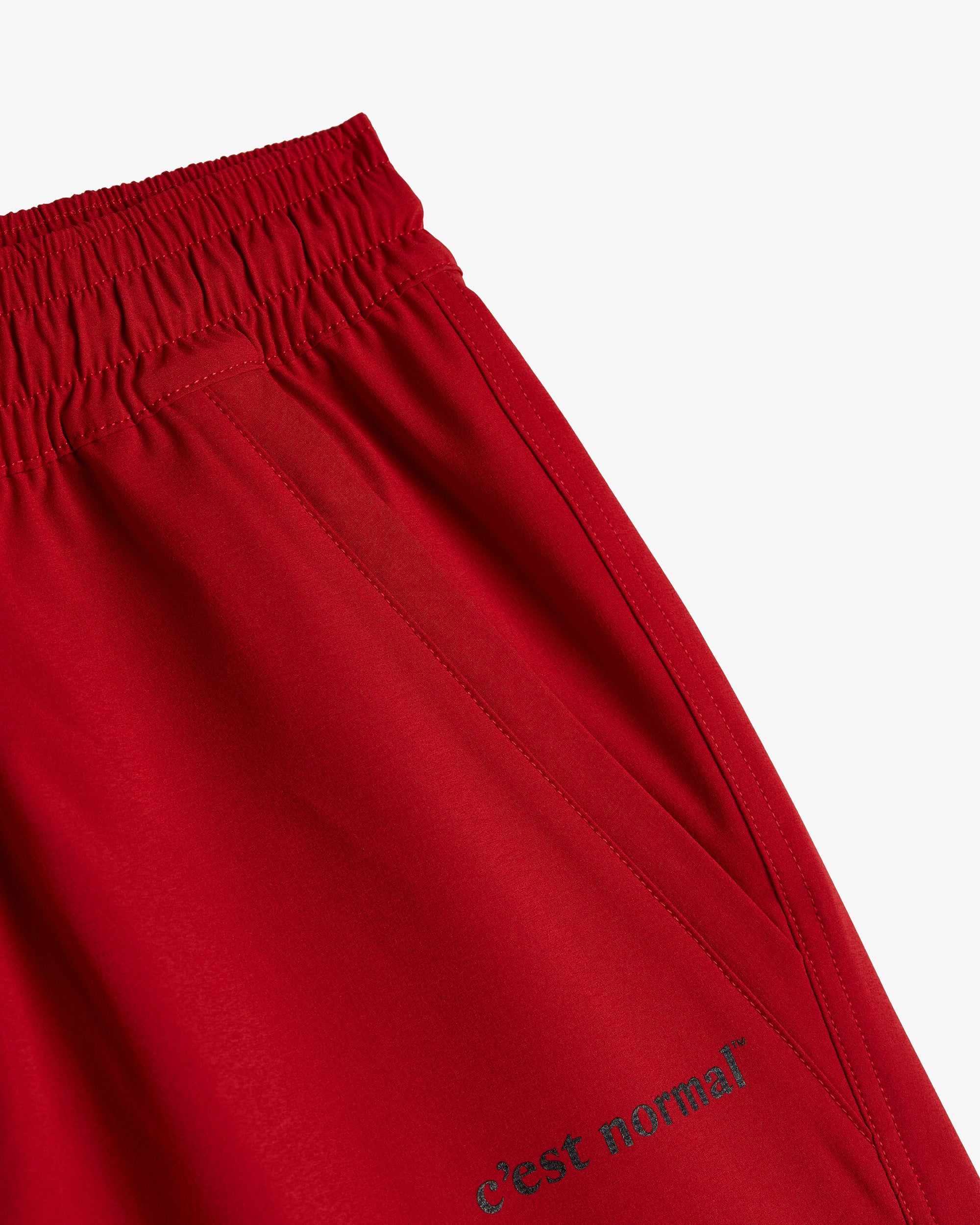 A detailed close-up of The Shorts in red, focusing on the elasticated waistband, side pocket, and the dark c'est normal logo on the leg.