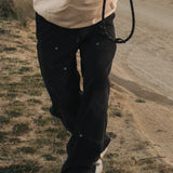 A close-up shot of a person wearing The Carpenter Pant in black, featuring visible copper-toned rivets, standing outdoors on a dirt path with one leg crossed over the other.