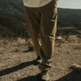 A close-up shot of a person wearing The Carpenter Pant in olive green, standing on a dirt path with a mountainous landscape in the background. The image highlights the straight leg fit, slash pockets, and copper-toned rivets of the pants.