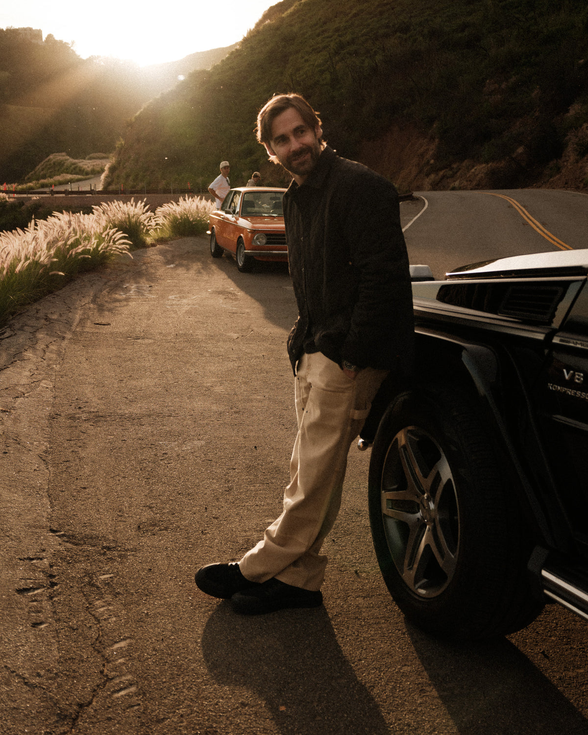 Warner wearing The Carpenter Pant in beige leans against a black car on a sunny road, looking to the side. The image highlights the relaxed, straight-leg fit and classic design of the pants by c'est normal.
