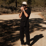 A man wearing The Carpenter Pant in black, a black t-shirt, and a c'est normal cap stands on a dirt road, holding a vintage camera up to his face. The background features a landscape with trees and distant hills.