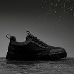 A side profile view of The Dynamic Waterproof Sneaker 2.0 Low in black waterproof. The shoe is sitting on a wet surface with water droplets falling on and around it against a grey background, showcasing its waterproof capabilities.