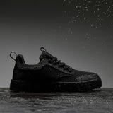 A side profile view of The Dynamic Waterproof Sneaker 2.0 Low in black waterproof. The shoe is sitting on a wet surface with water droplets falling on and around it against a grey background, showcasing its waterproof capabilities.