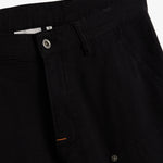 A detailed close-up of The Carpenter Pant in black, highlighting the front button closure, textured fabric, and the embroidered c'est normal logo near the pocket.