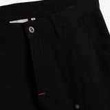 A detailed close-up of The Carpenter Pant in black, highlighting the front button closure, textured fabric, and the embroidered c'est normal logo near the pocket.