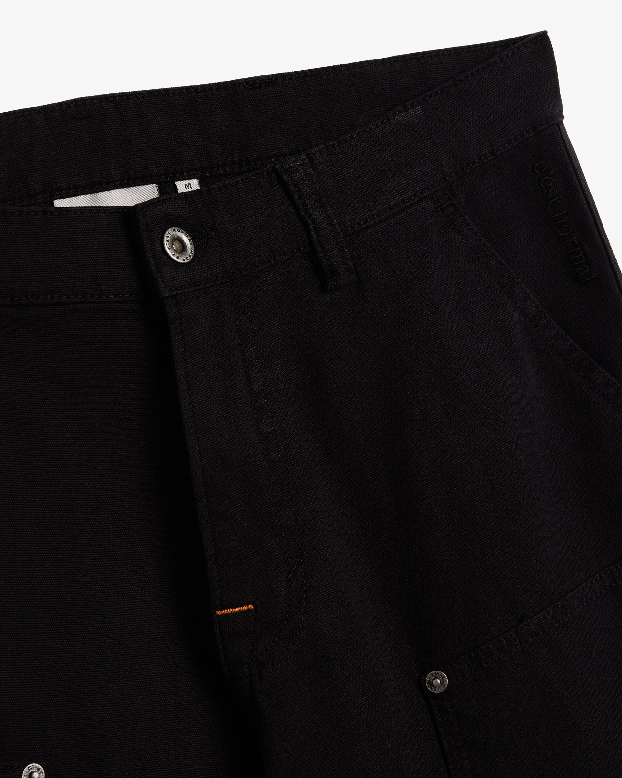 A detailed close-up of The Carpenter Pant in black, highlighting the front button closure, textured fabric, and the embroidered c'est normal logo near the pocket.