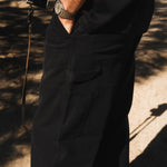 A close-up view of a person wearing The Carpenter Pant in black, with their hand in the front pocket. The image highlights the textured cotton fabric, side tool pockets, and hammer loop, characteristic of classic workwear.