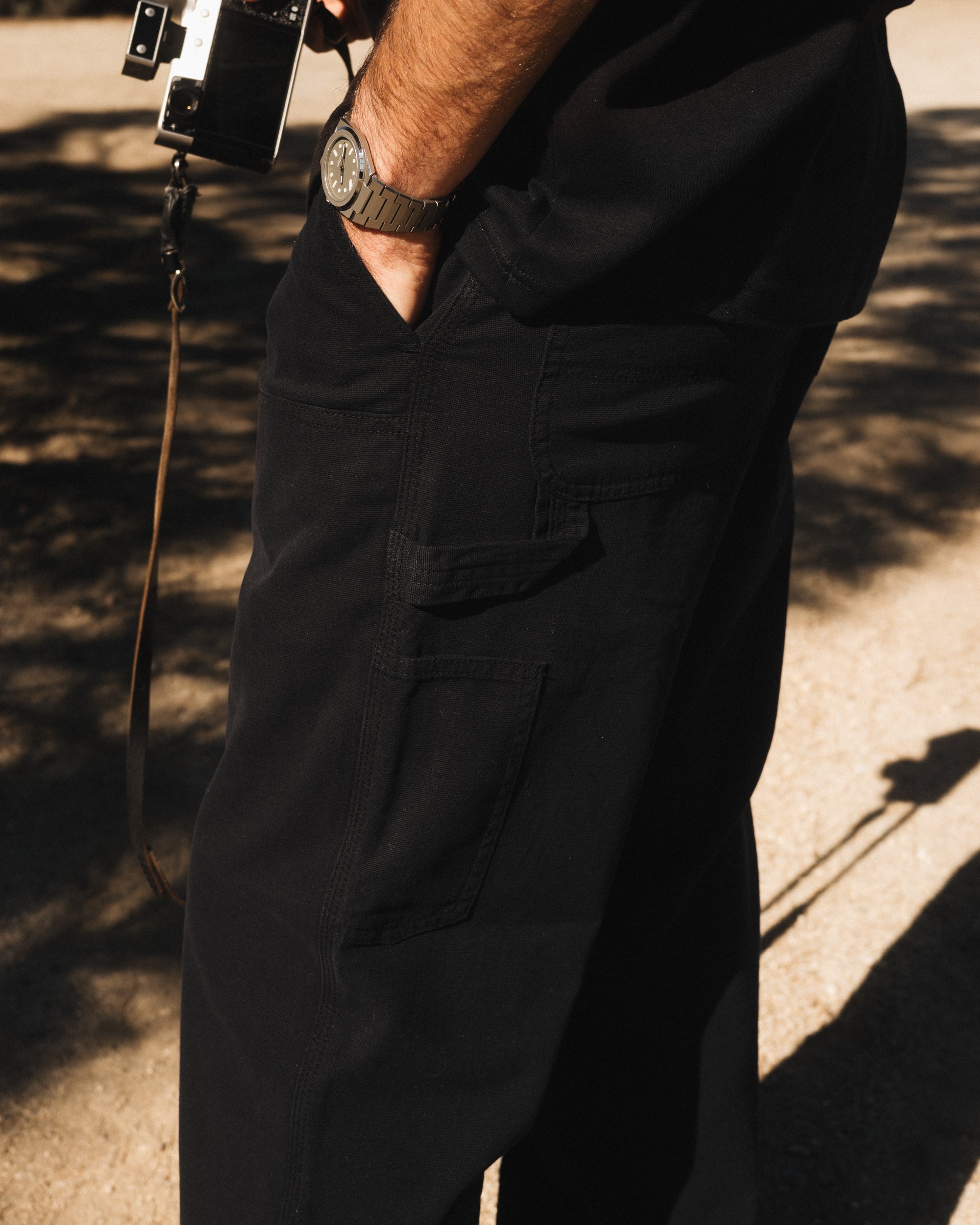 A close-up view of a person wearing The Carpenter Pant in black, with their hand in the front pocket. The image highlights the textured cotton fabric, side tool pockets, and hammer loop, characteristic of classic workwear.