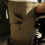 A close-up shot of the side pockets on The Carpenter Pant in olive green, showing the textured fabric and pocket details. The person is standing next to a motorcycle, wearing a white t-shirt.