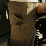 A close-up shot of the side pockets on The Carpenter Pant in olive green, showing the textured fabric and pocket details. The person is standing next to a motorcycle, wearing a white t-shirt.