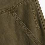 A close-up view of The Carpenter Pant in olive green, focusing on the textured fabric and pocket detailing. The image clearly shows the stitched c'est normal logo near the pocket seam.