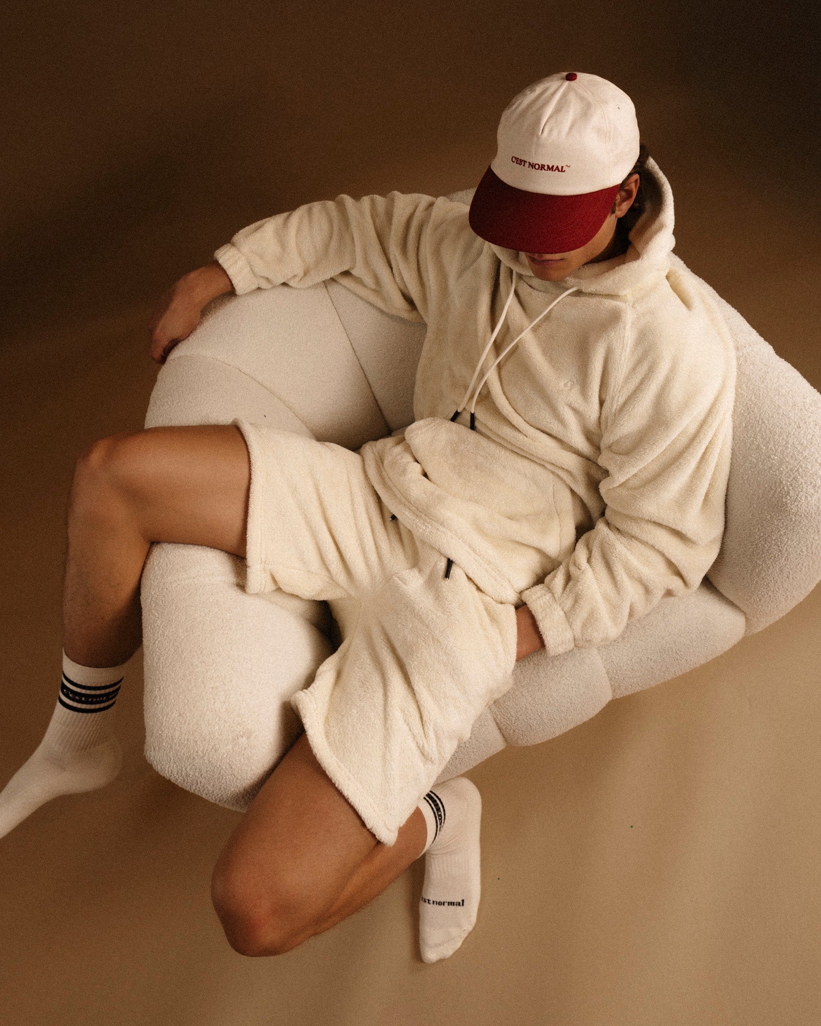 The Oversized Teddy Hoodie in cream — worn with matching shorts in a white textured chair