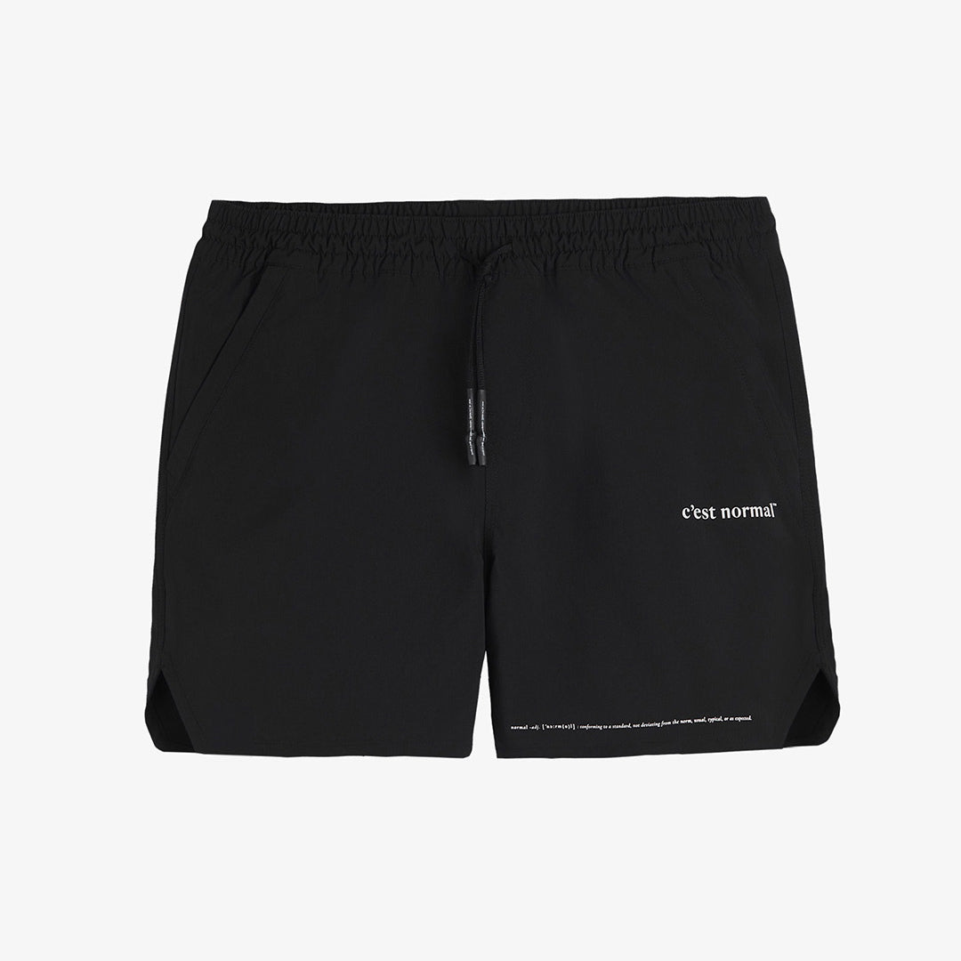 A front view of The Shorts in black, laid flat on a white background. The image highlights the elastic waistband with drawstrings, zippered side pockets, notched hem, and the white c'est normal logo on the left leg.