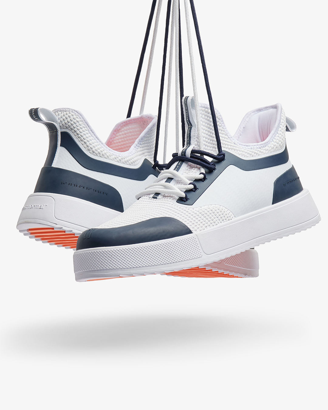A pair of The Sneakers 2.0 in white and navy, suspended in the air by their dark navy laces. The image highlights the white mesh upper, navy blue rubber accents, thick white EVA midsole, and the c'est normal logo on the heel, revealing the original orange rubber grid on the sole.