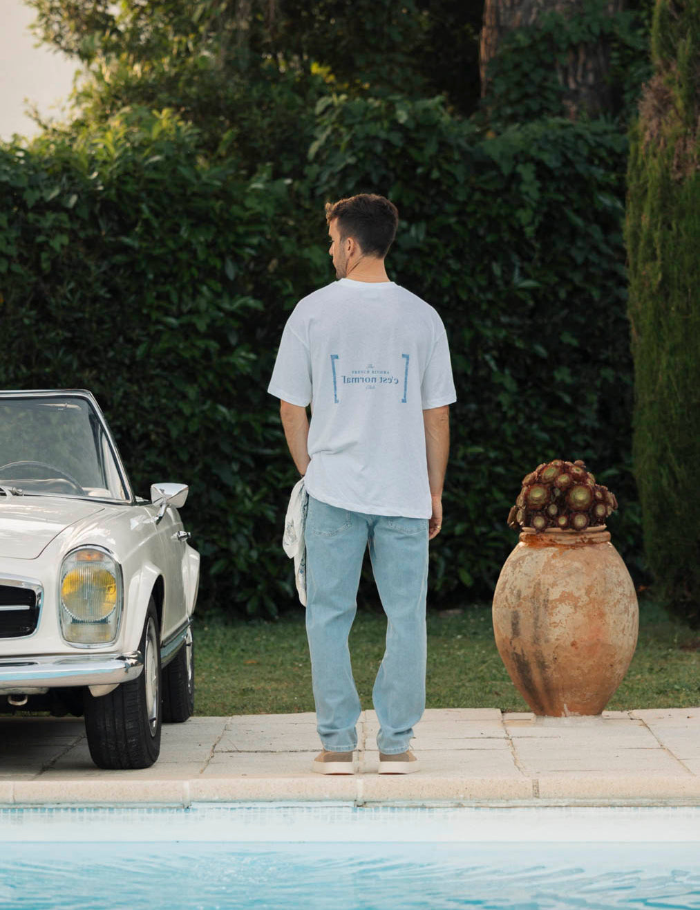 Le Linen T-Shirt in white — back view with C'est normal graphic next to a pool and vintage car