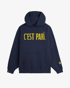 A front view of The c'est pari Oversized Hoodie in navy, laid flat on a white background. The image highlights the large yellow c'est pari print on the chest, a kangaroo pocket, and a c'est normal cuff tag on the right sleeve.