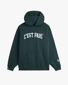 A front view of The c'est pari Oversized Hoodie in pine, laid flat on a white background. The image highlights the white c'est pari logo on the chest, a kangaroo pocket, and the c'est normal tag on the right cuff.