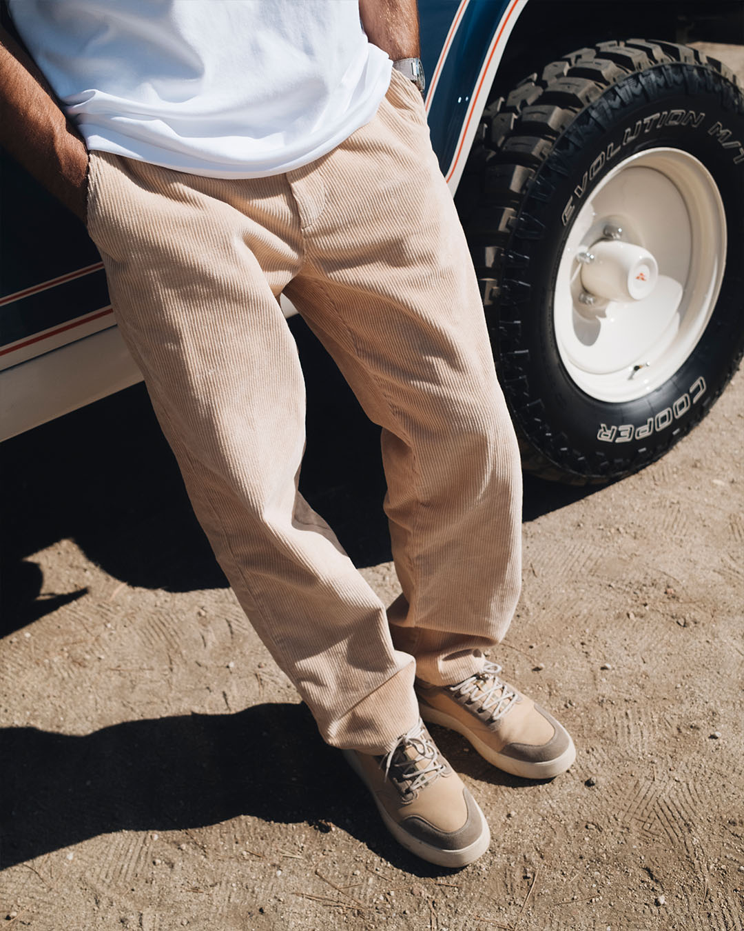 A close-up shot of a person leaning against a vehicle outdoors, wearing The CÔTELÉ Corduroy Straight Pants in Irish Cream paired with a white t-shirt and beige sneakers. The image highlights the straight leg fit and textured mid wale corduroy fabric.