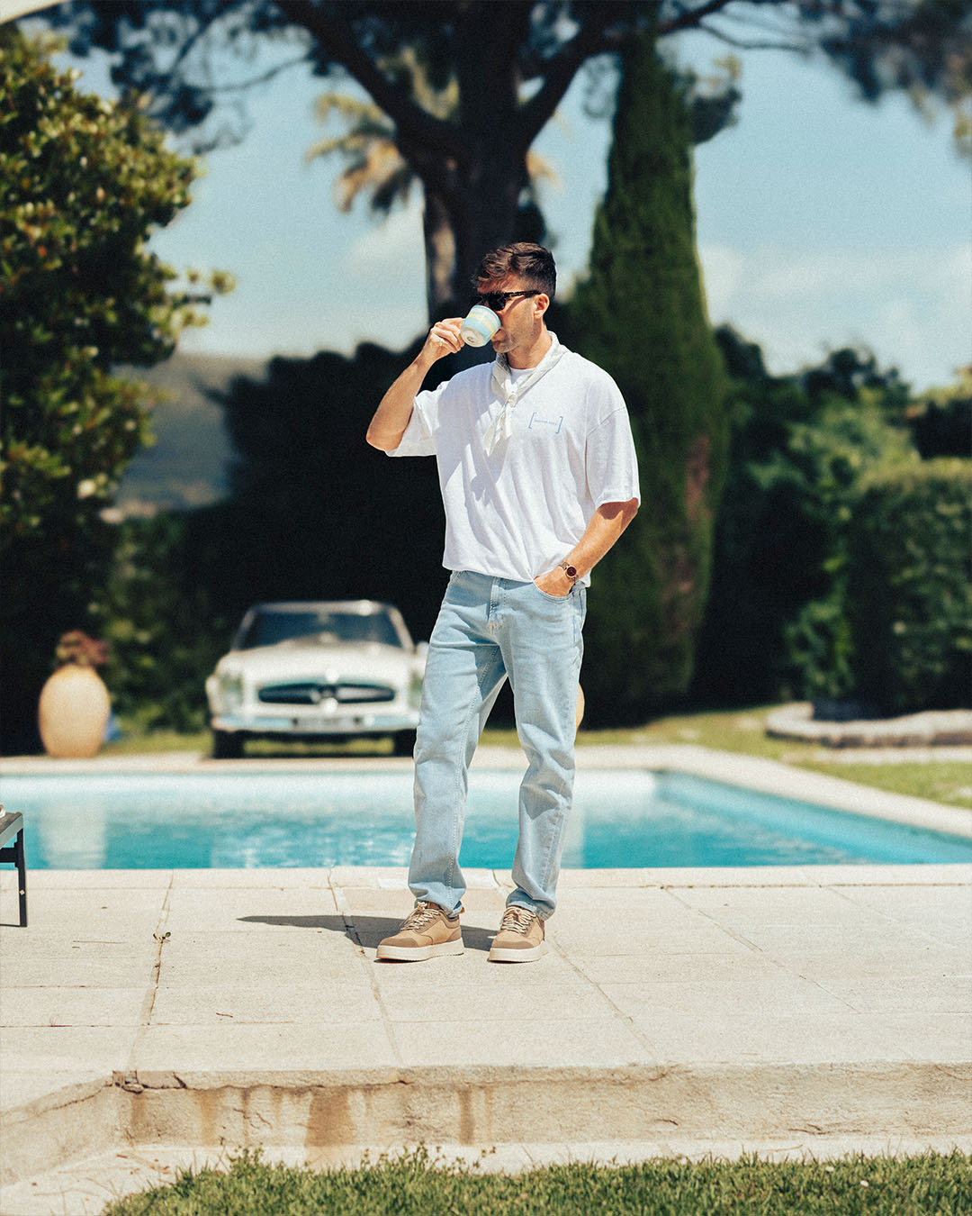 A man wearing The Jeans Relaxed Straight Fit in light blue by c'est normal, along with a white t-shirt and sunglasses, stands by a swimming pool drinking from a mug. A classic white car is visible in the background.