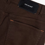 A close-up shot of the back of The 5 Pocket Pants in brown, focusing on the back pocket and the black rubber patch on the waistband featuring the white c'est normal logo and orange accent stitching.