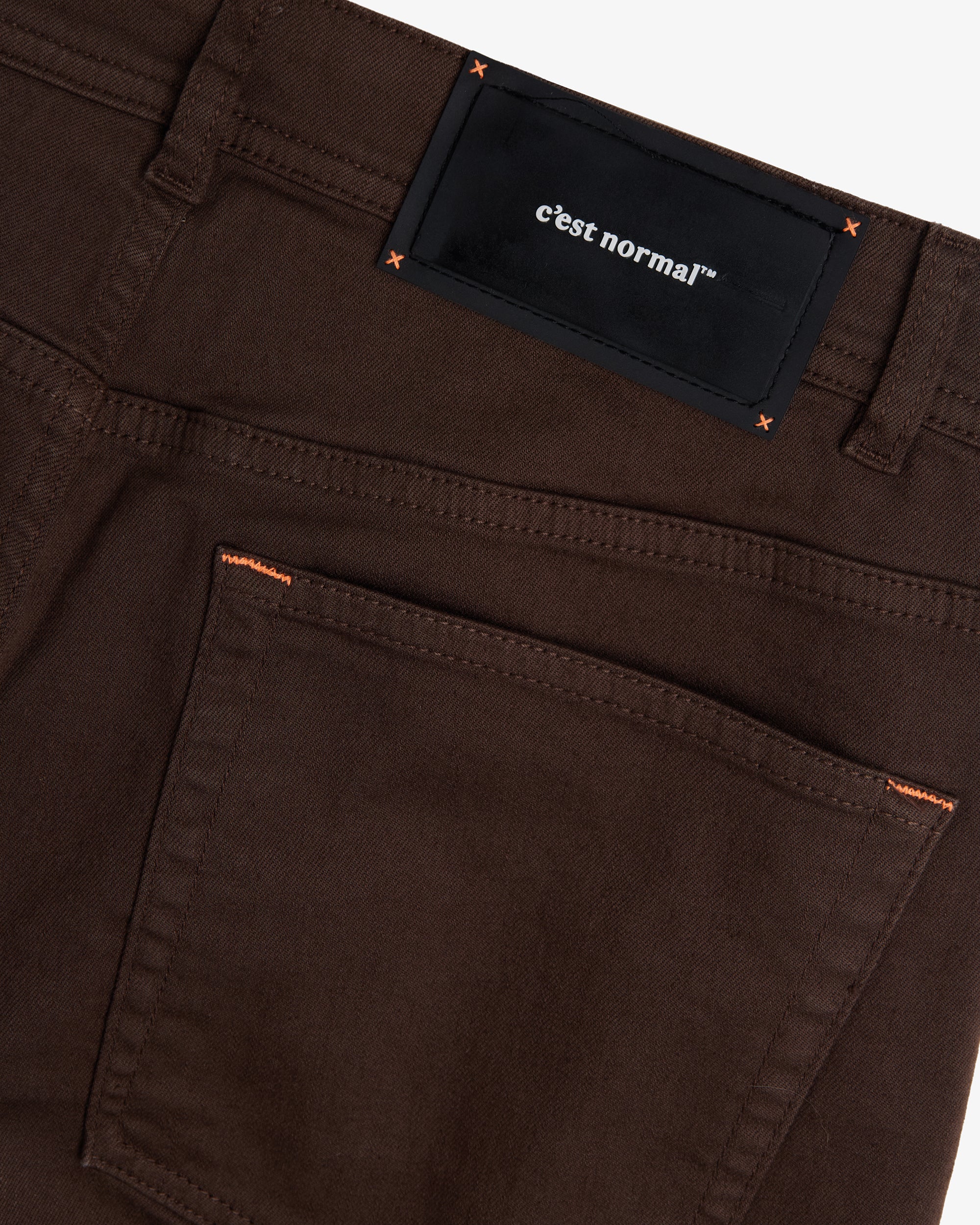 A close-up shot of the back of The 5 Pocket Pants in brown, focusing on the back pocket and the black rubber patch on the waistband featuring the white c'est normal logo and orange accent stitching.