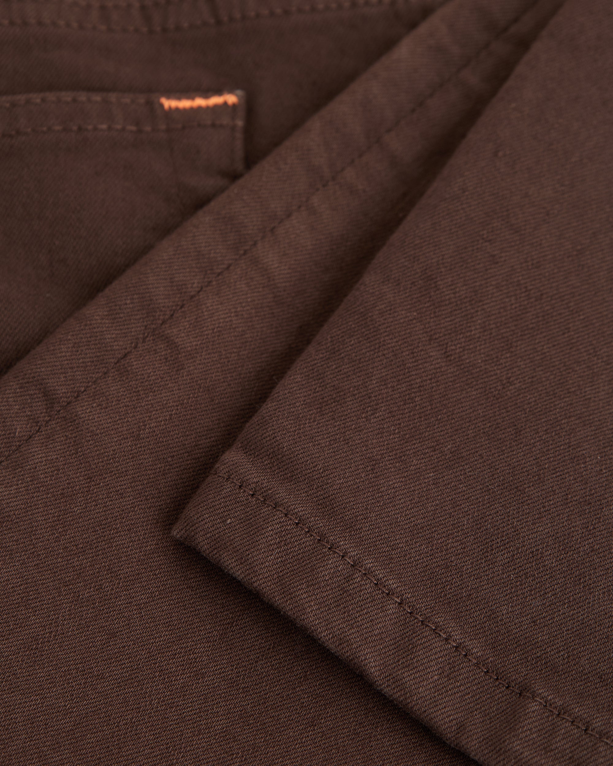 A close-up shot of the soft, stretchy denim fabric of The 5 Pocket Pants in brown, highlighting the texture and a touch of orange accent stitching on the pocket.