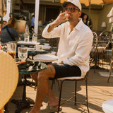 The Board Shorts in black — worn with a white linen shirt and cap while sitting at a sunlit cafe