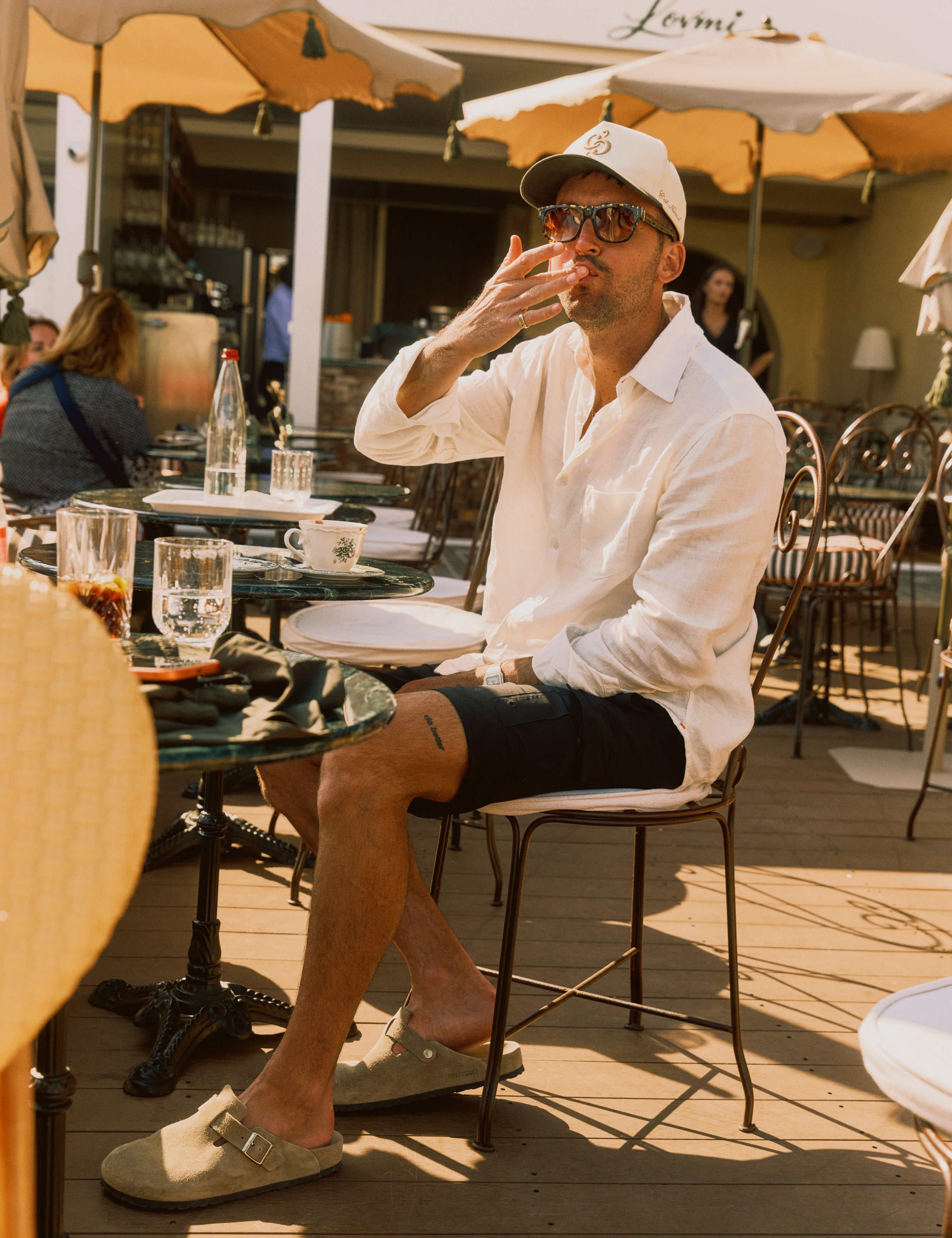 The Board Shorts in black — worn with a white linen shirt and cap while sitting at a sunlit cafe