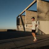 A man jumping rope outdoors against a concrete wall wearing The Athlétique Tee in sage green, paired with black shorts and a black c'est normal cap.