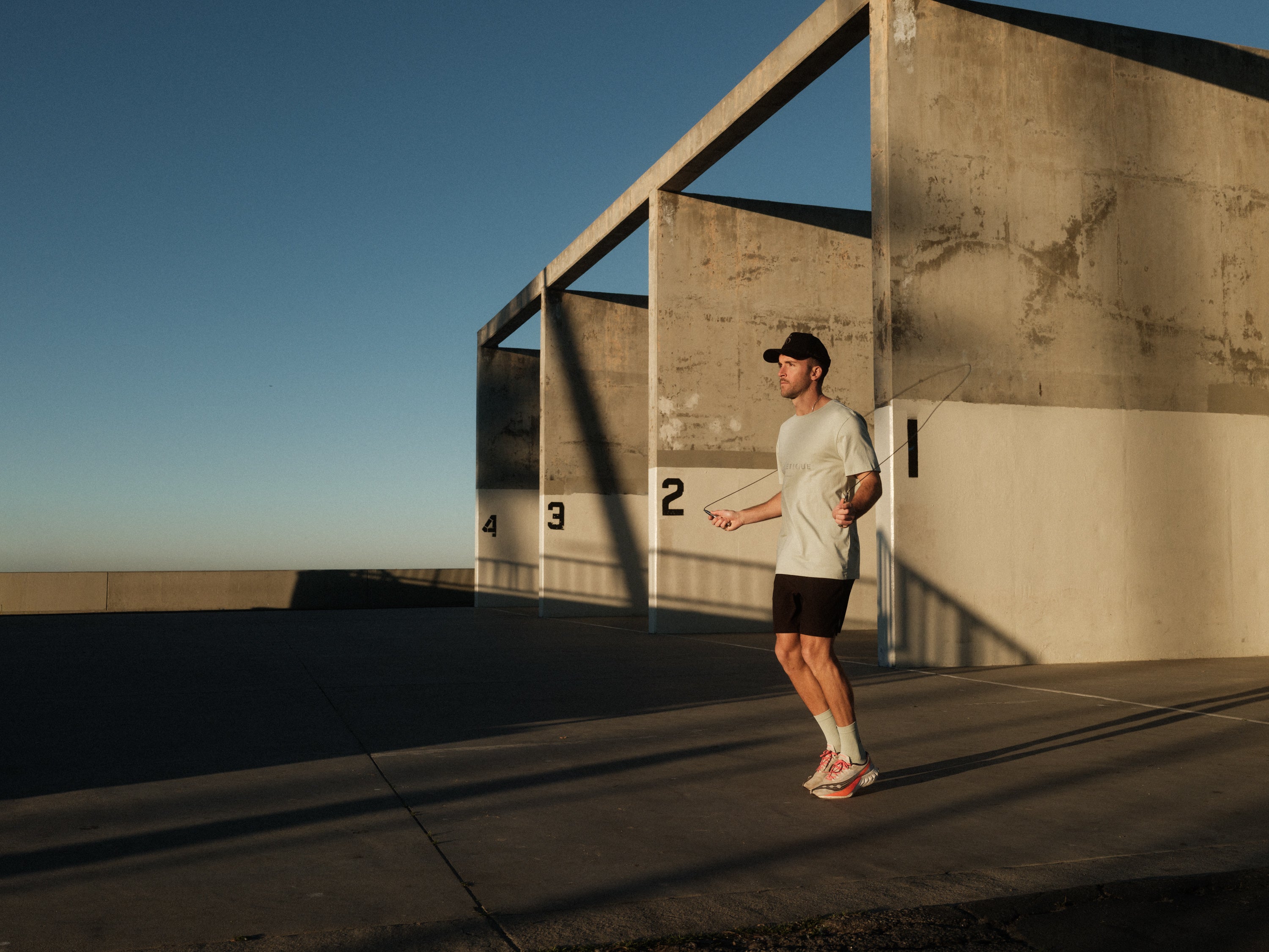 A man jumping rope outdoors against a concrete wall wearing The Athlétique Tee in sage green, paired with black shorts and a black c'est normal cap.