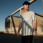A man wearing The Athlétique Tee in sage green and a black cap stands outdoors holding a jump rope. The image highlights the white ATHLÉTIQUE logo and c'est normal text on the chest.