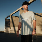 A man wearing The Athlétique Tee in sage green and a black cap stands outdoors holding a jump rope. The image highlights the white ATHLÉTIQUE logo and c'est normal text on the chest.