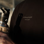A close-up shot of a person wearing The Oversized Gym T-Shirt in black, holding a water bottle. The image highlights the Athlétique logo and c'est normal text on the center front.