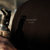 A close-up shot of a person wearing The Oversized Gym T-Shirt in black, holding a water bottle. The image highlights the Athlétique logo and c'est normal text on the center front.