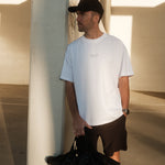 A man stands outdoors leaning against a concrete pillar, wearing The Oversized Gym T-Shirt in white. The shirt features the Athlétique and c'est normal logos on the center front. He is also wearing black shorts and a black cap, and holding a black duffel bag.
