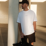 A man stands outdoors leaning against a concrete pillar, wearing The Oversized Gym T-Shirt in white. The shirt features the Athlétique and c'est normal logos on the center front. He is also wearing black shorts and a black cap, and holding a black duffel bag.