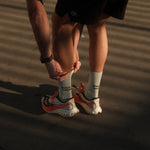 A person wearing The Athlétique Socks in sage green, paired with running shoes and black shorts, bends down to adjust them. The image highlights the mid-calf length and the black ATHLÉTIQUE and c'est normal text printed on the side.