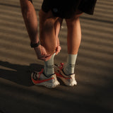 A person wearing The Athlétique Socks in sage green, paired with running shoes and black shorts, bends down to adjust them. The image highlights the mid-calf length and the black ATHLÉTIQUE and c'est normal text printed on the side.