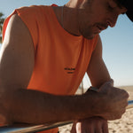 A close-up of a man wearing The Athlétique Tank in orange and a black cap, leaning on a metal bar outdoors. The image highlights the small black ATHLÉTIQUE and c'est normal logo on the chest.
