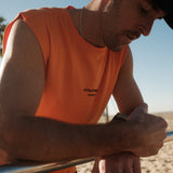 A close-up of a man wearing The Athlétique Tank in orange and a black cap, leaning on a metal bar outdoors. The image highlights the small black ATHLÉTIQUE and c'est normal logo on the chest.