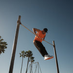 A man wearing The Athlétique Tank in orange, black pants, and a black c'est normal cap, performs a muscle-up on an outdoor bar against a backdrop of palm trees and a clear blue sky.