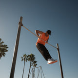A man wearing The Athlétique Tank in orange, black pants, and a black c'est normal cap, performs a muscle-up on an outdoor bar against a backdrop of palm trees and a clear blue sky.