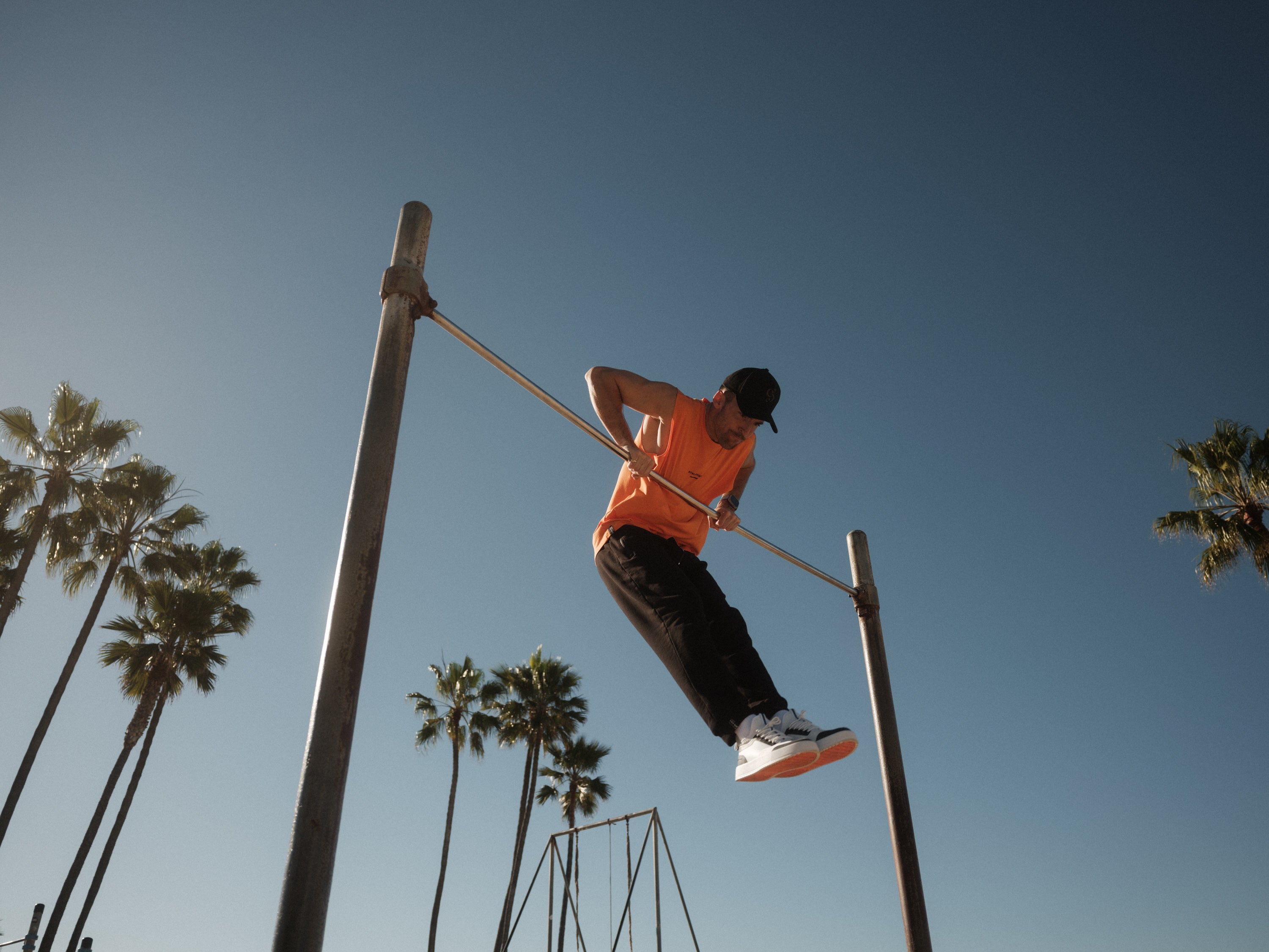 A man wearing The Athlétique Tank in orange, black pants, and a black c'est normal cap, performs a muscle-up on an outdoor bar against a backdrop of palm trees and a clear blue sky.