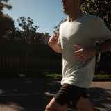 A man runs outdoors wearing The Athlétique Tee in sage green, paired with black shorts, a black cap, and sunglasses. The image highlights the relaxed fit of the t-shirt and the Athlétique logo on the chest.