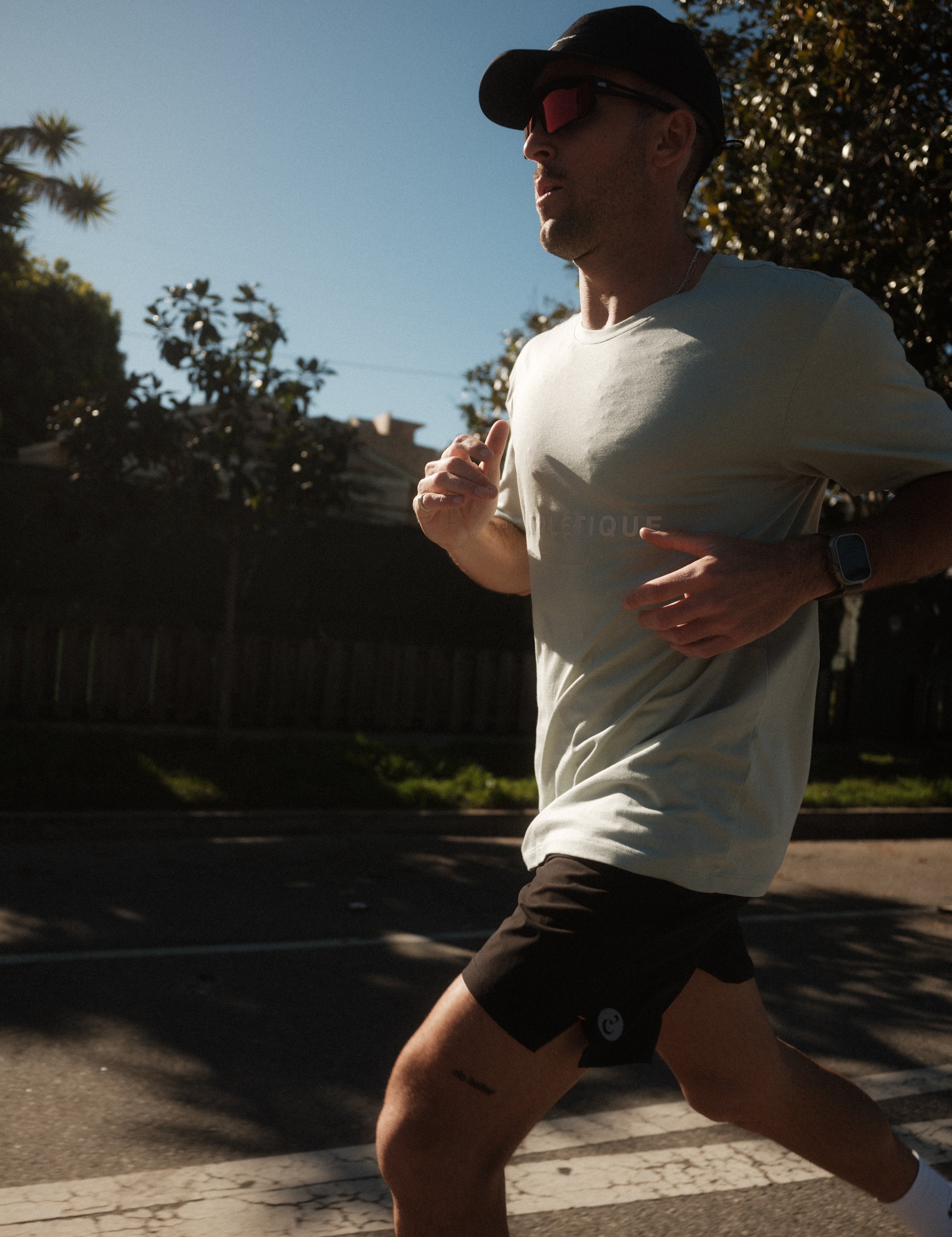 A man runs outdoors wearing The Athlétique Tee in sage green, paired with black shorts, a black cap, and sunglasses. The image highlights the relaxed fit of the t-shirt and the Athlétique logo on the chest.