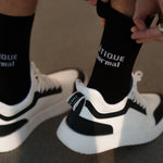 A person pulling up a pair of The Athlétique Socks in black, featuring the c'est normal logo in contrast stitching. They are also wearing black and white sneakers.