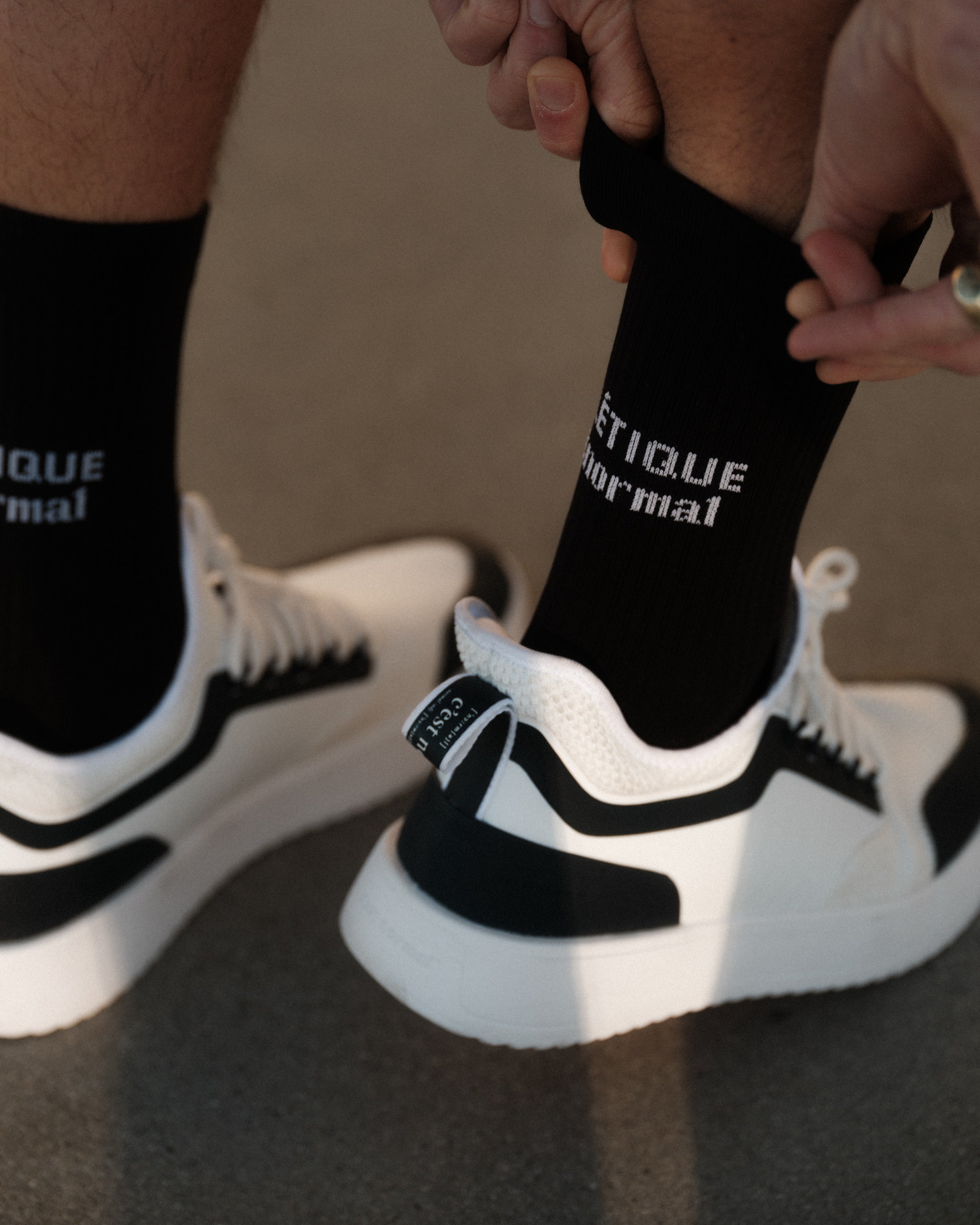 A person pulling up a pair of The Athlétique Socks in black, featuring the c'est normal logo in contrast stitching. They are also wearing black and white sneakers.
